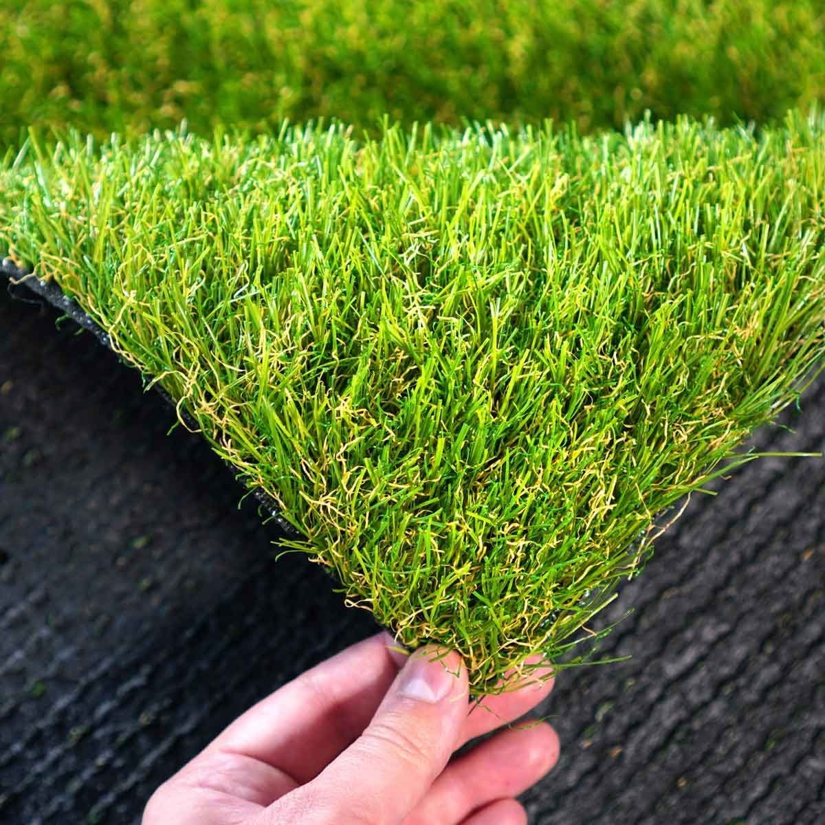 A hand holds a piece of artificial grass, displaying its vivid green blades, against a textured dark surface in the background.