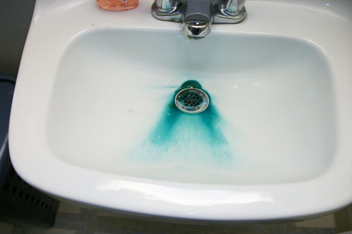 A white sink contains water with a blue-green discoloration surrounding the drain, suggesting a possible cleaning agent or dye. Soap is near