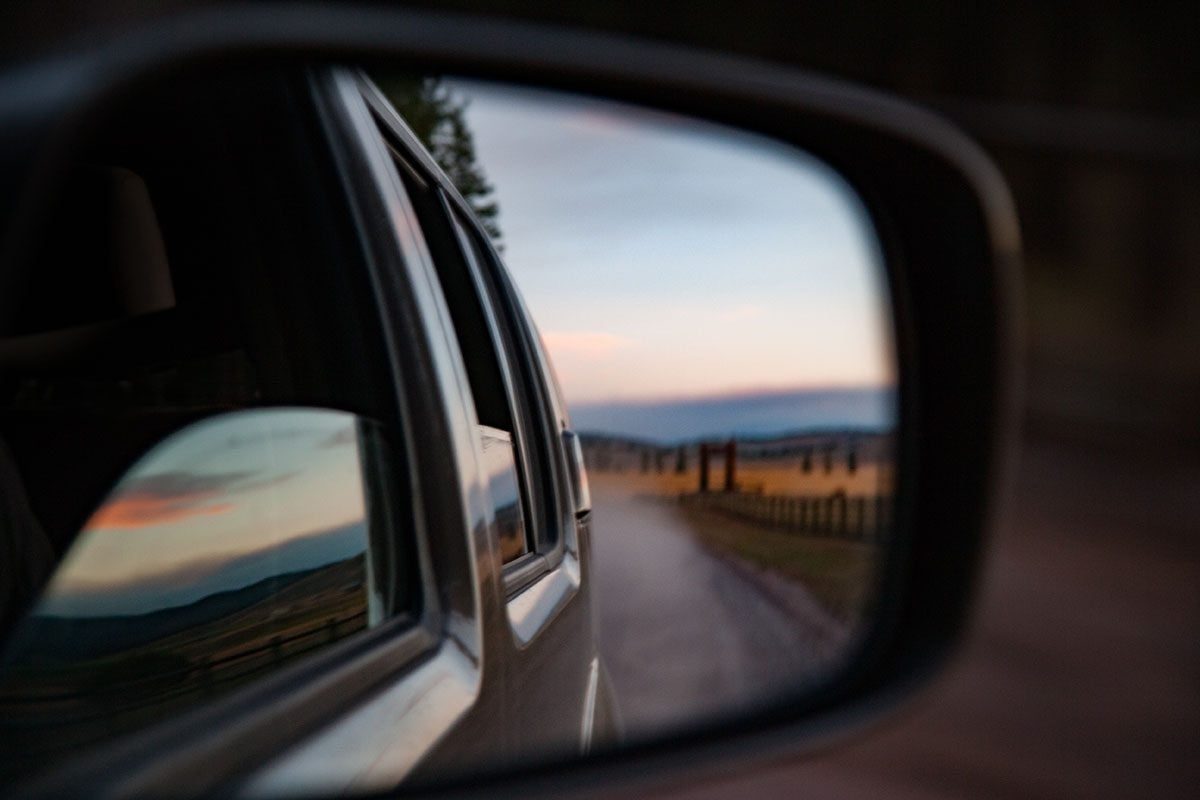 A side mirror reflects a winding road with distant mountains and a sunset, while a vehicle drives away from the scene.