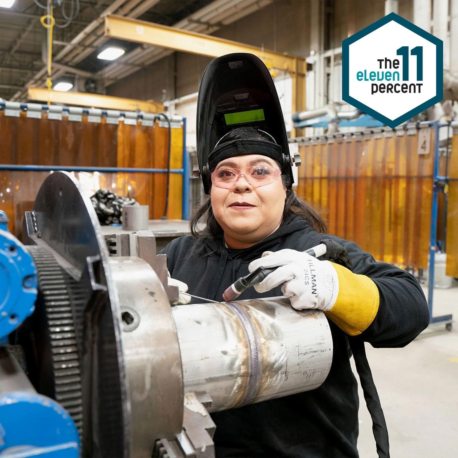 A welder, wearing protective gear, is working with metal machinery in a workshop, focusing on welding a cylindrical piece against a backdrop