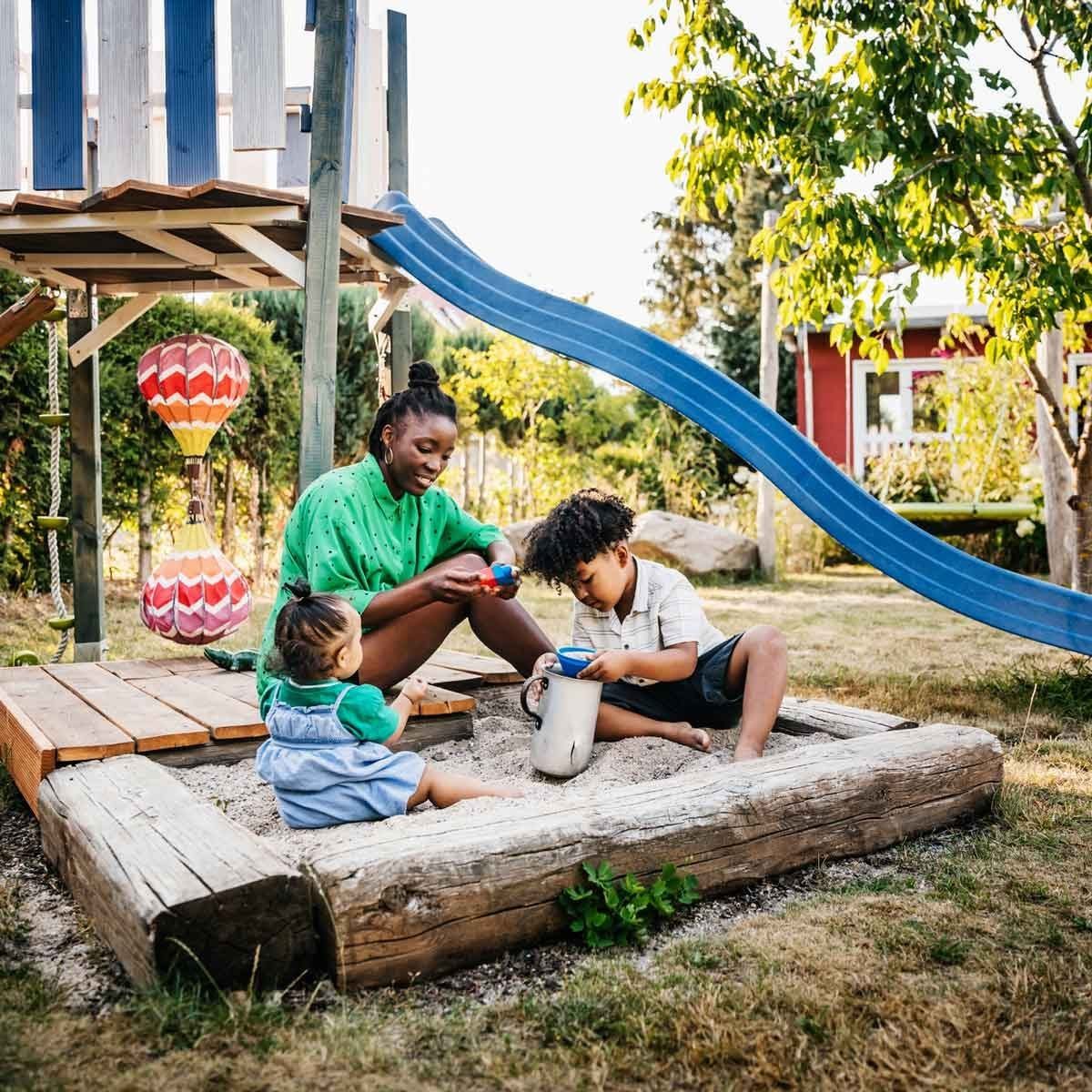 A woman and two children play in a sandpit, using toys. A blue slide and greenery surround them in a vibrant, outdoor playground setting.