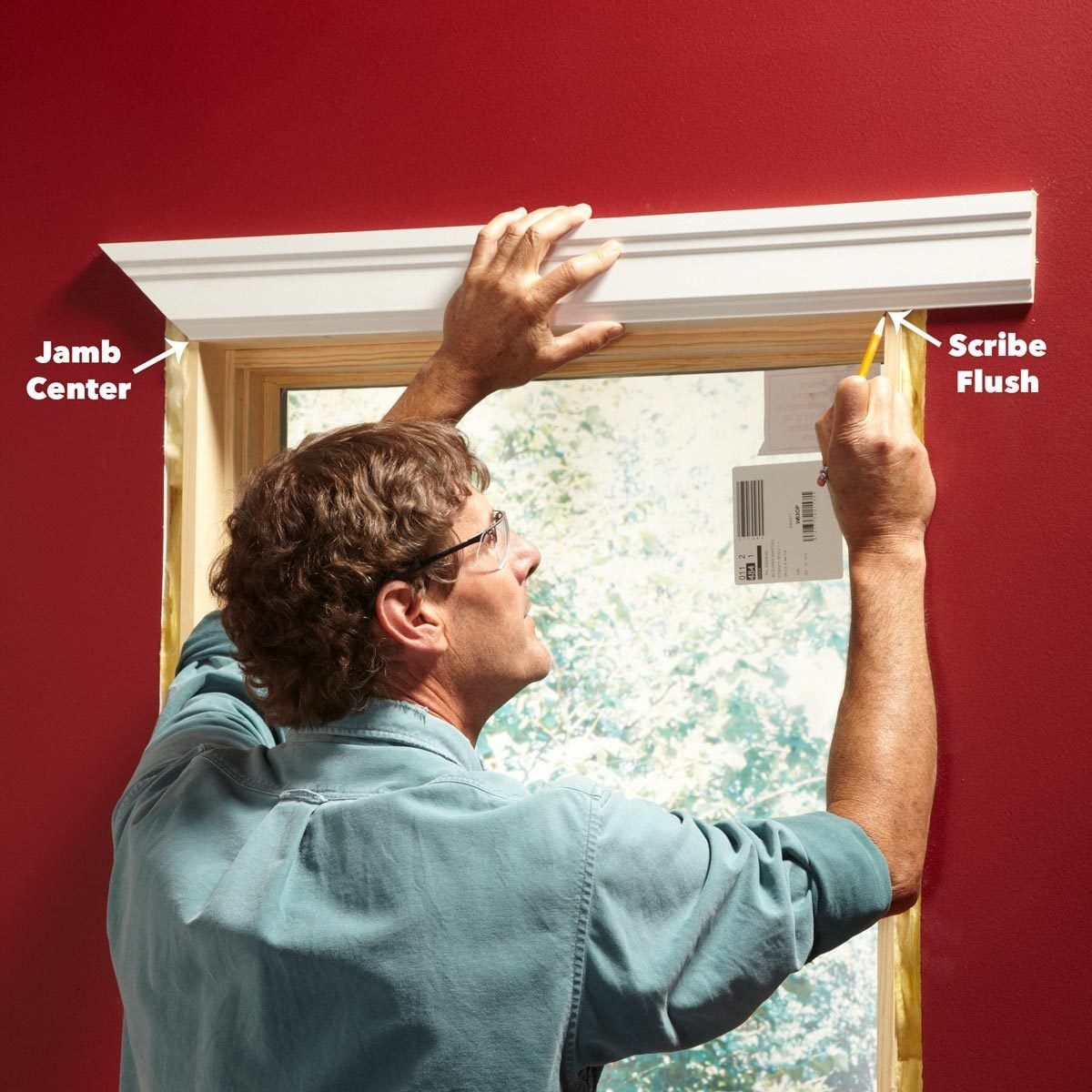 A man measures and marks a wooden trim above a window, surrounded by a red wall and natural light from the window.