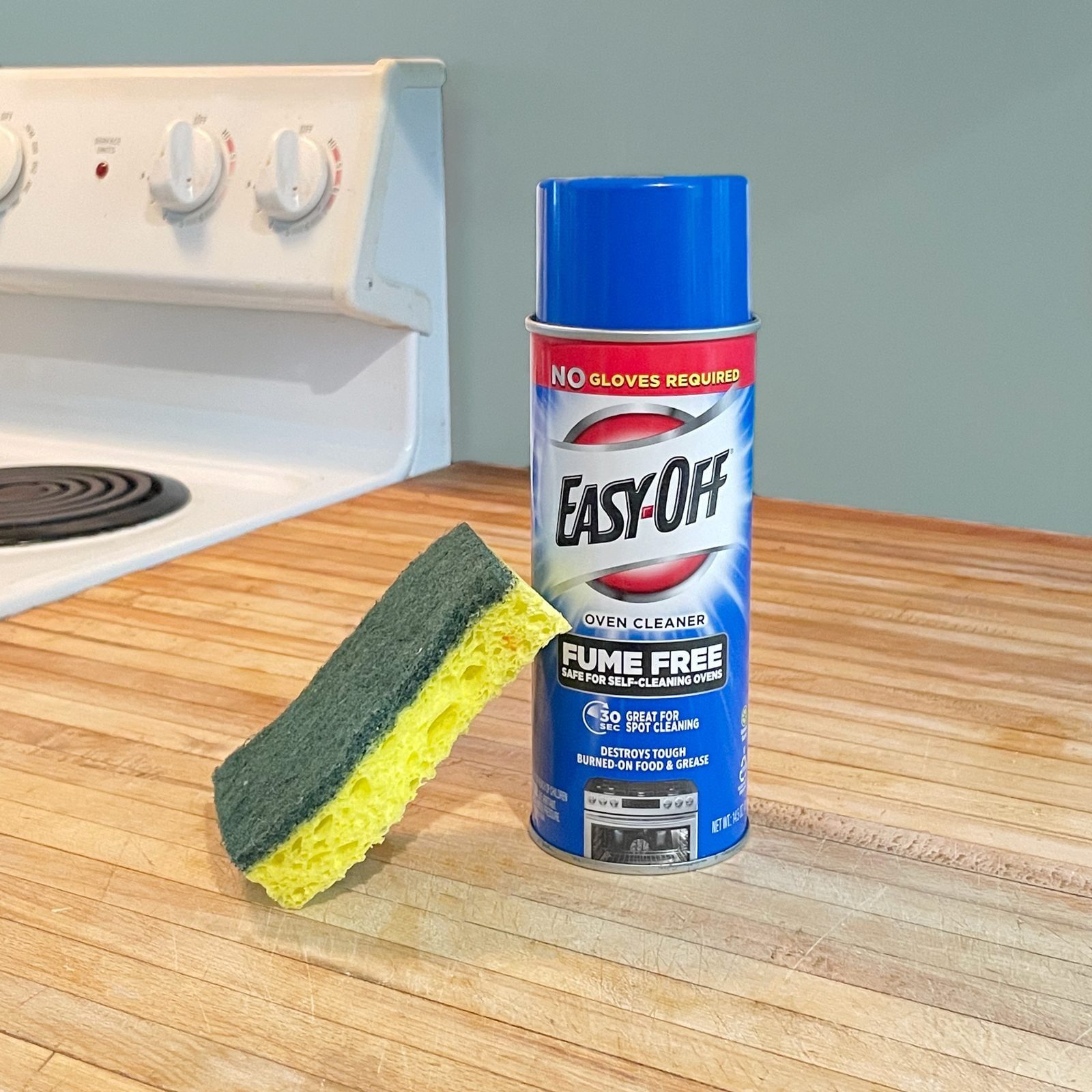 A can of Easy-Off oven cleaner stands next to a yellow-and-green sponge on a wooden countertop near a white stove in a kitchen.