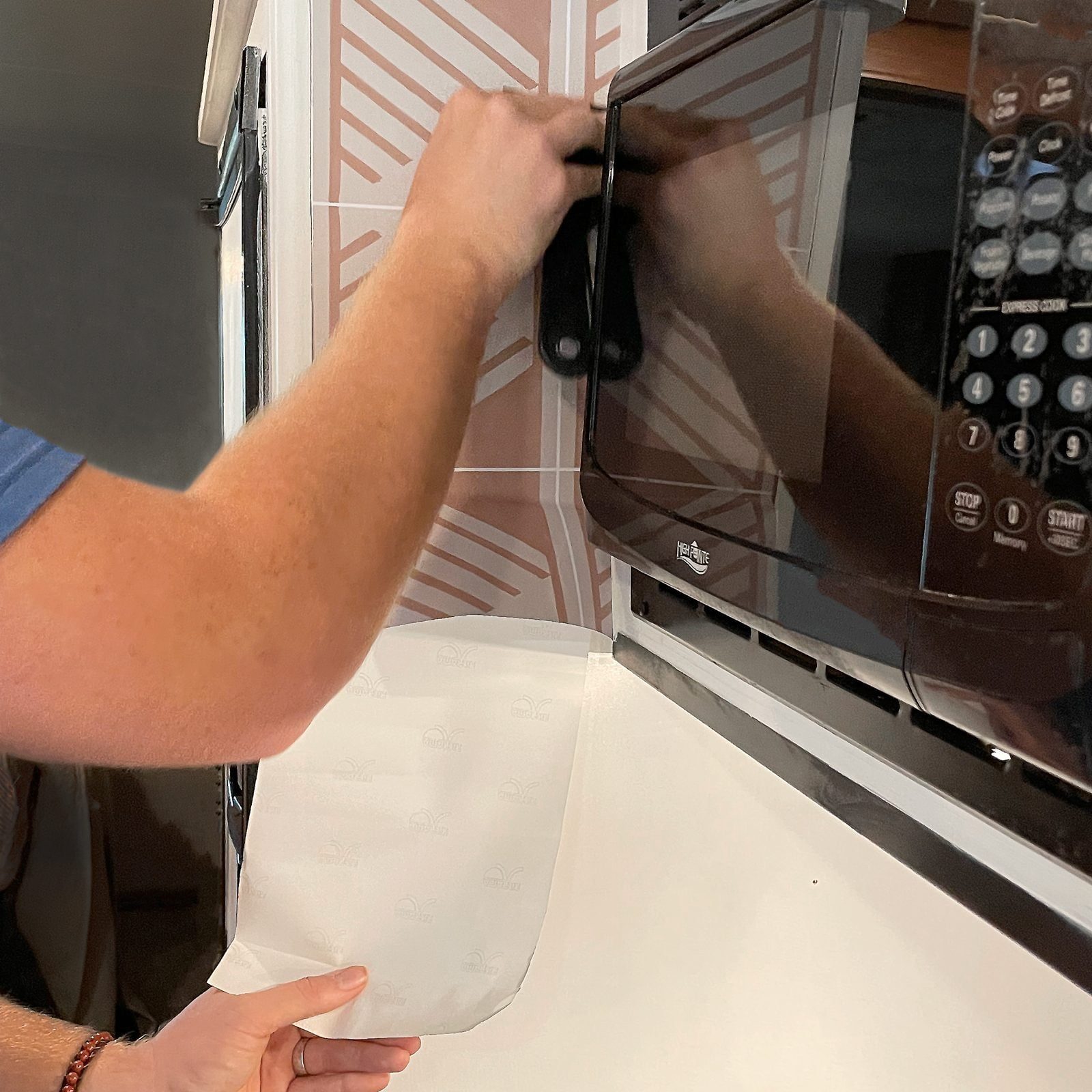 A person holds a piece of paper while adjusting a microwave's installation against a patterned wall in a kitchen setting.