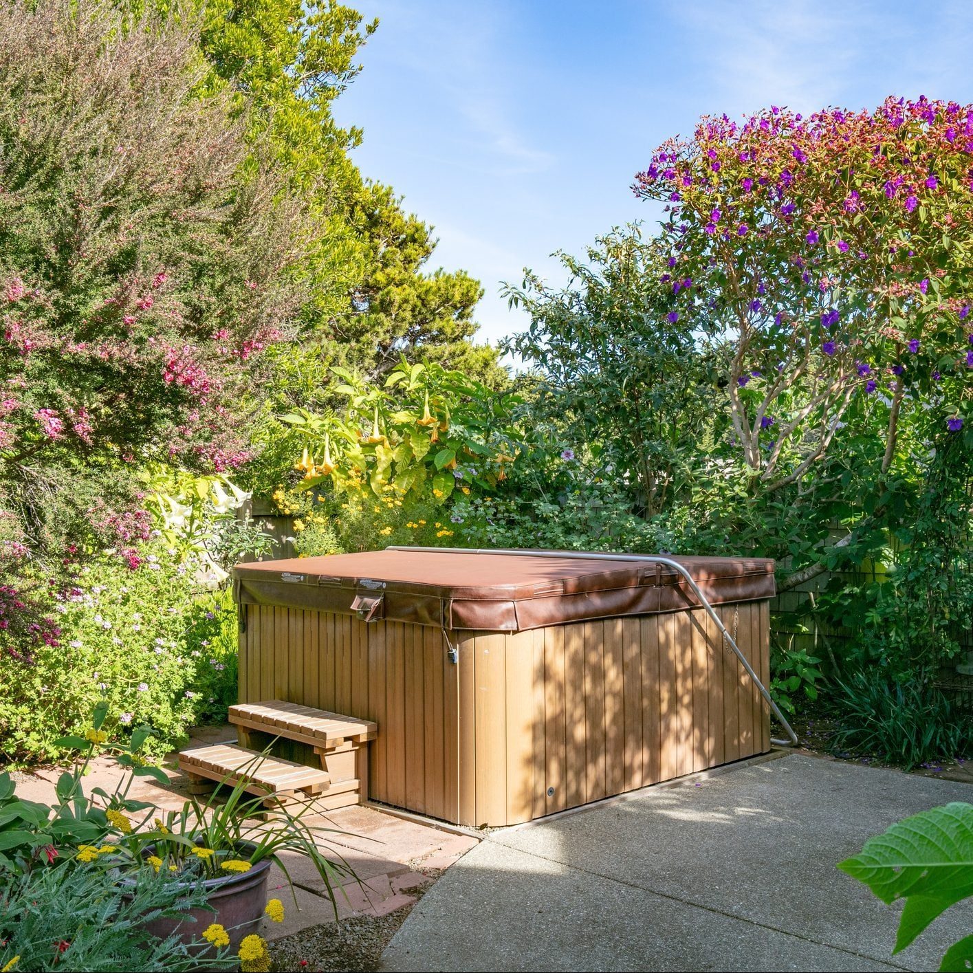 A hot tub sits among lush greenery and colorful flowers, with wooden steps leading to its side, surrounded by a well-maintained garden path.