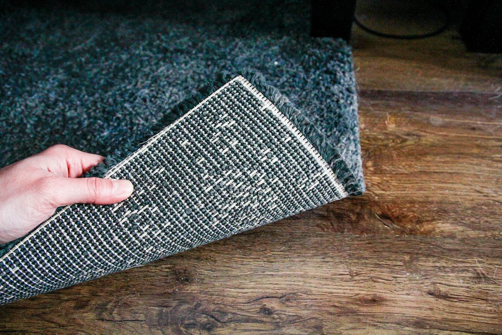 A hand is lifting a corner of a patterned carpet, revealing the underside, against a wooden floor backdrop.
