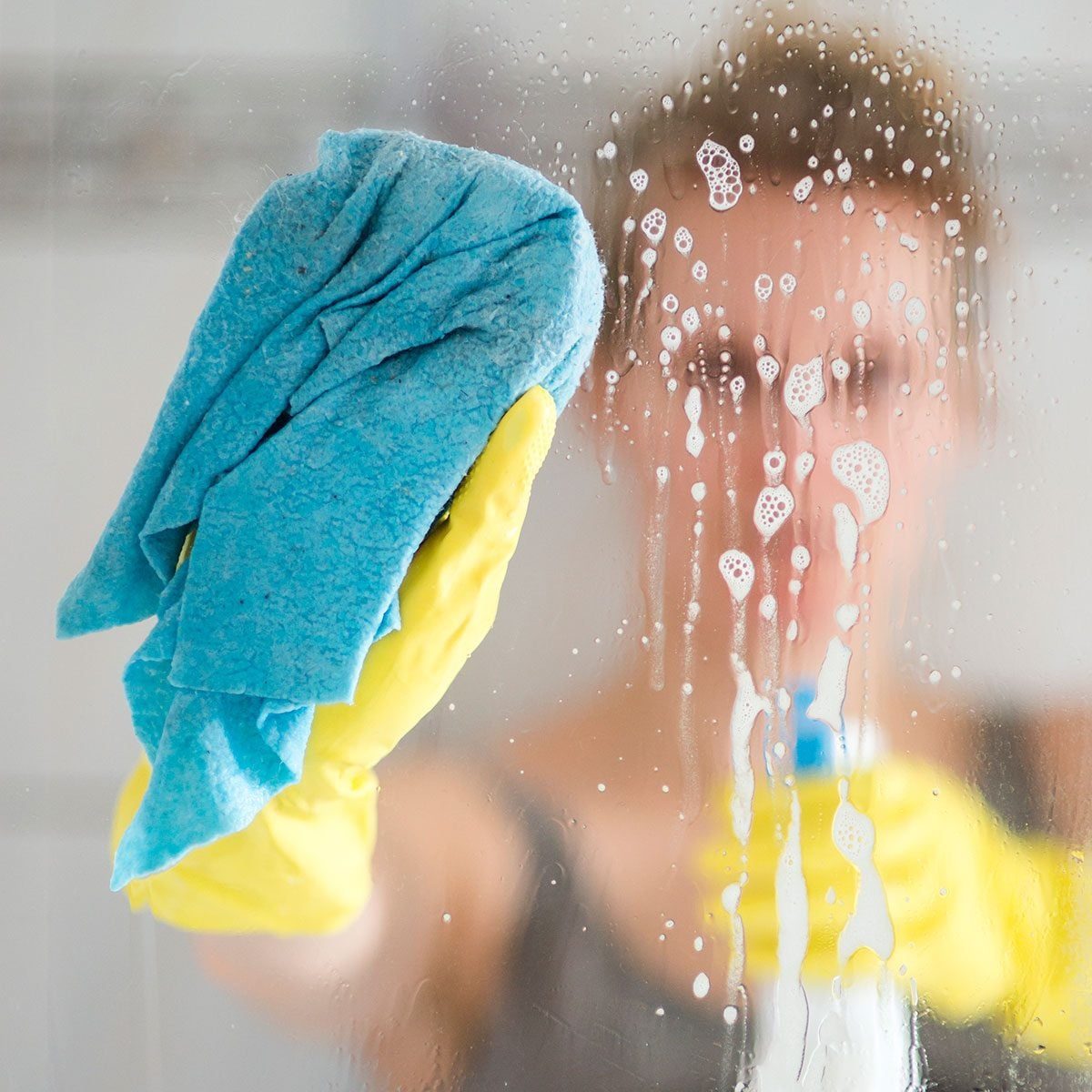 A gloved hand wipes a soapy cloth across a foggy glass surface, revealing a blurred figure holding a spray bottle in a bathroom setting.