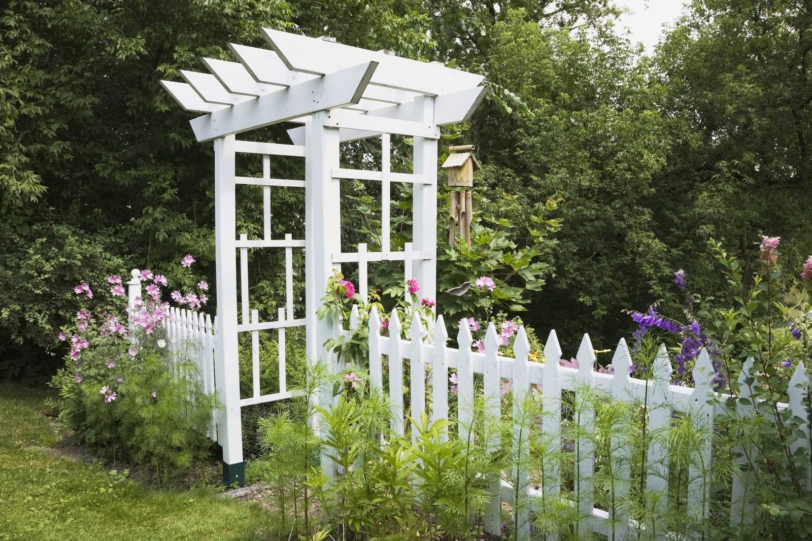 A white garden arbor stands surrounded by colorful flowers and a picket fence, set against a lush green backdrop of trees.
