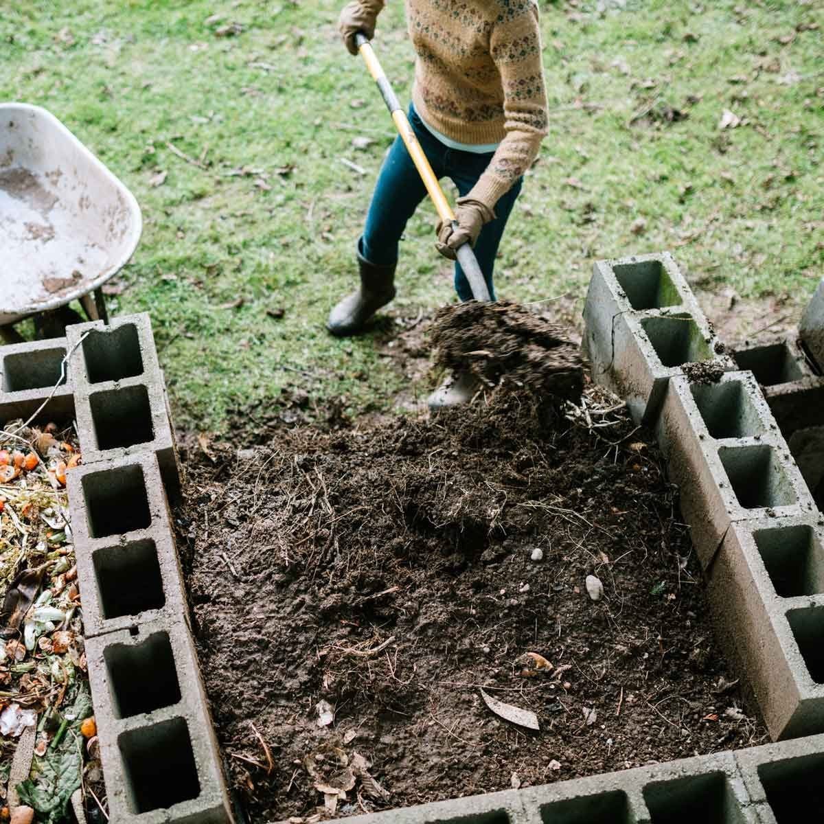 A person shovels compost into a block structure in a grassy outdoor area, surrounded by earthy materials and a wheelbarrow in the background